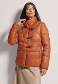 Bruine pufferjas met een hoge kraag, zwarte togglesluiting, quilted ontwerp en een gladde textuur. Draagt een beige rok.