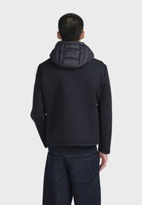 Felpa nera con cappuccio imbottito, caratterizzata da una texture liscia e una vestibilità casual. Abbinata a jeans in denim scuro.