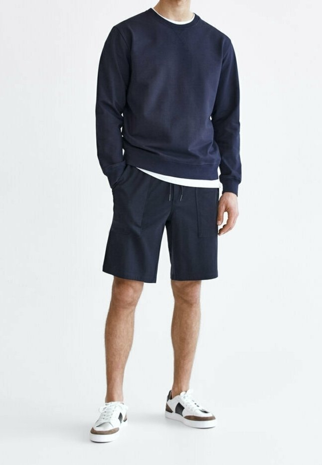 Sweat-shirt en coton marine à manches longues et col rond, associé à un short foncé avec des poches avant, porté avec des baskets blanches et noires.