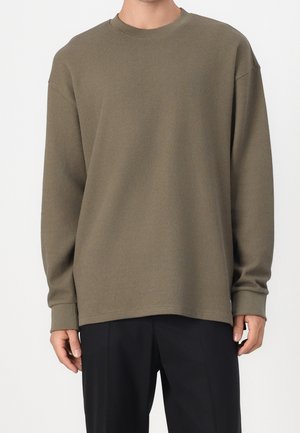 Homme portant un pull côtelé olive à manches longues et un pantalon noir, debout devant un fond blanc uni.