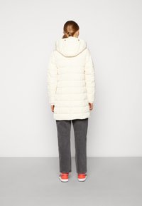 Abrigo largo tipo puffer acolchado en color crema con capucha removible. Presenta un corte recto y puños acanalados; se lleva sobre pantalones gris oscuro y zapatillas.