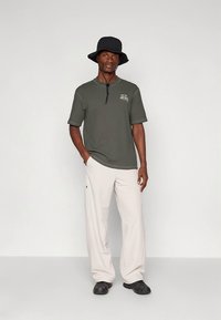 Short-sleeve olive green ribbed shirt with collar, logo detail; beige loose fitting pants; black bucket hat; black shoes. Simple, casual outfit.