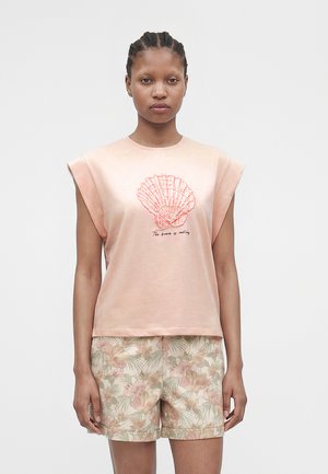 Vrouw met een mouwloze perzikkleurige top met een rode schelp en tekst, gecombineerd met beige bloemenprint shorts, staand tegen een effen lichte achtergrond.