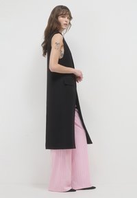 Schwarzer ärmelloser langer Schal mit geradem Schnitt, versehen mit einem seitlichen Schlitz und einer Tasche, kombiniert mit rosa gestreiften weit geschnittenen Hosen.