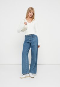 Hvid langærmet top med en rund halsudskæring, parret med højhævede, brede blå jeans. Accessorierne inkluderer en lille, lyse farvet håndtaske.