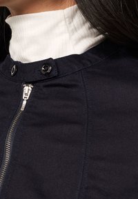 Giacca blu navy con chiusura a zip e bottoni a scatto, caratterizzata da un alto colletto a coste bianco e una texture del tessuto liscia.