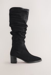 Schwarzer Wildleder-Mittelschaftstiefel mit gestapeltem Blockabsatz und lockerem Schaft vor hellem einfarbigem Hintergrund.