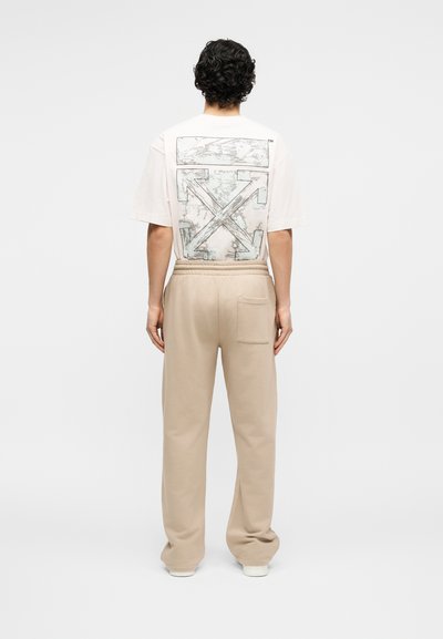 Άνδρας με λευκό φαρδύ μπλουζάκι με μαύρο αφηρημένο σχέδιο βέλους και μπεζ φαρδιά παντελόνια, στέκεται γυρισμένος με την πλάτη σε απλό λευκό φόντο.