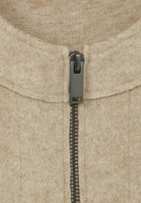 Close-up van een beige wollen kledingstuk met een zwart metalen rits die gedeeltelijk geopend is in het midden.