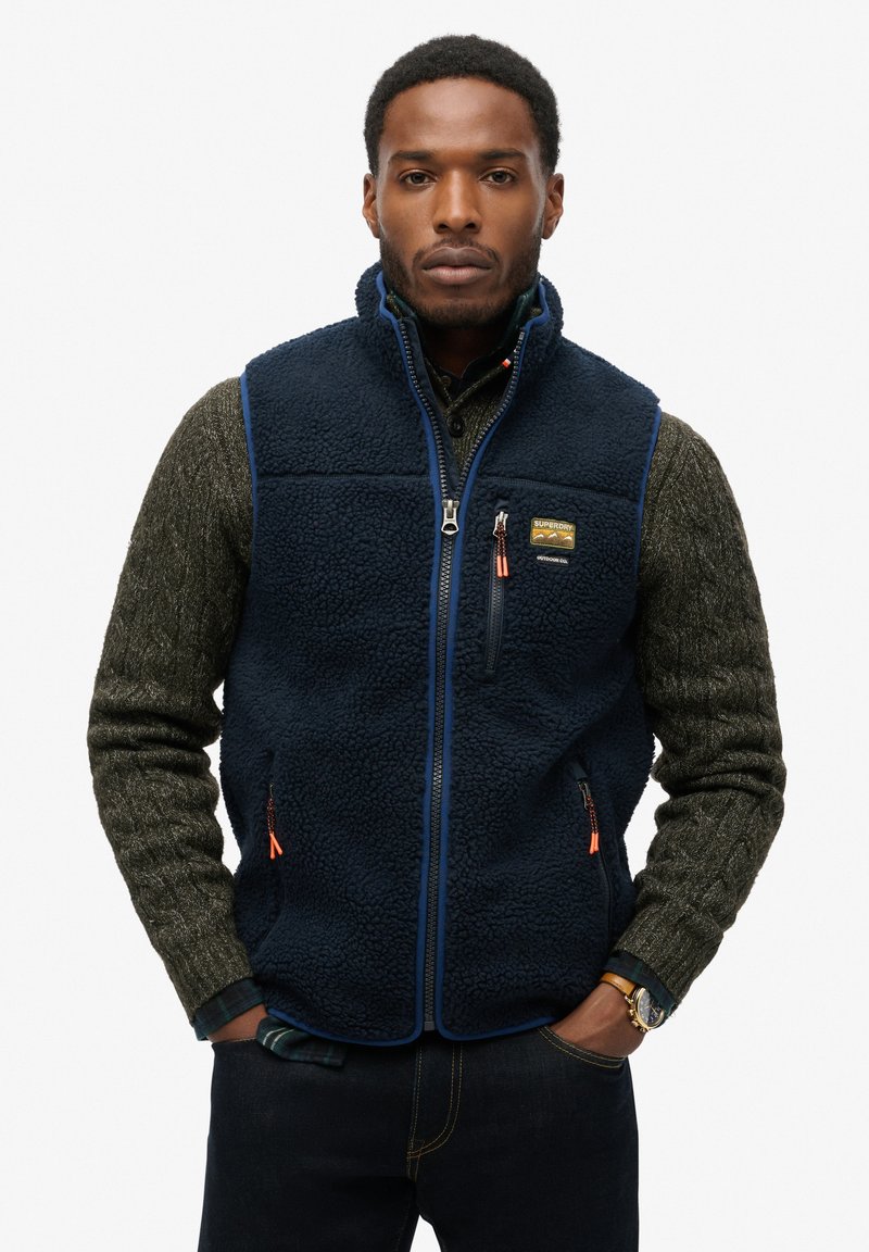 Homme portant une veste en polaire bleu marine sur un pull texturé vert olive, les mains dans les poches et une montre-bracelet au poignet gauche.