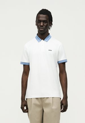 Άνδρας που φοράει άσπρο πόλο μπλουζάκι με ανοικτό μπλε γιακά και άκρες στα μανίκια, μπεζ παντελόνι, στέκεται μπροστά σε απλό ανοιχτό φόντο.