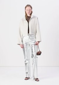 Lyhyeksi leikattu vaalea harmaa takki, jossa on leveät hihat ja etutaskut, yhdistettynä kiiltäviin hopeisiin suoriin housuihin ja mustiin korkokenkiin.