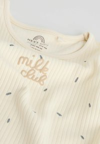 Chemise bébé côtelée crème avec texte "milk club" et petites formes de feuilles grises éparpillées, montrant l'encolure et l'étiquette de la marque à l'intérieur du col.