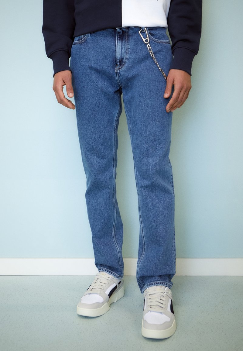 Jeans de mezclilla de color azul claro con un corte recto, que presentan un detalle de cadena plateada en el bolsillo frontal, combinados con zapatillas blancas y negras.