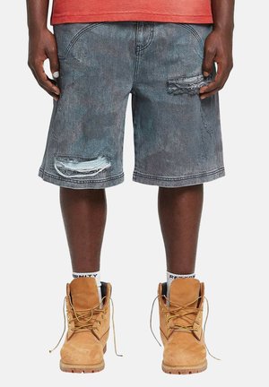 Person trägt ausgewaschene schwarze Denim-Shorts mit Abnutzungen, braune Schnürstiefel und weiße Socken mit schwarzem Text.