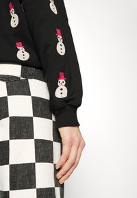 Maglione nero con stampe di pupazzi di neve e cappelli rosa; abbinato a pantaloni a scacchi bianchi e neri. Mano visibile appoggiata sui pantaloni.