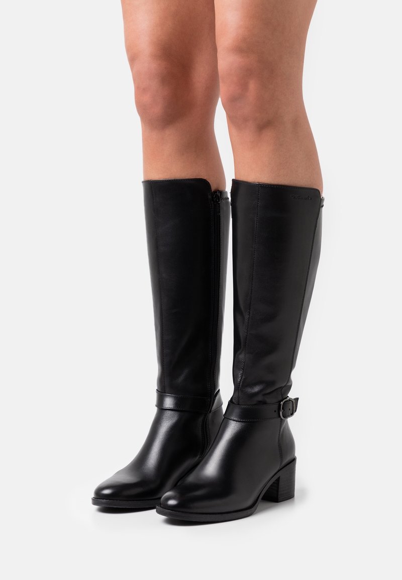 Jambes portant des bottes en cuir noir montantes avec talons blocs et fermetures éclair latérales, debout sur un fond blanc.