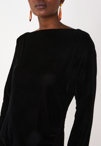 Robe longue en velours noir à manches longues avec un décolleté carré et des détails froncés sur le côté. Les accessoires comprennent des boucles d'oreilles géométriques orange.