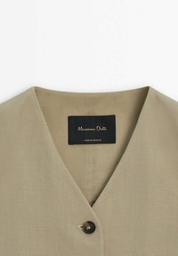 Vêtement beige à col en V avec un bouton foncé unique et une étiquette noire à l'intérieur mentionnant Massimo Dutti, fabriqué au Portugal.