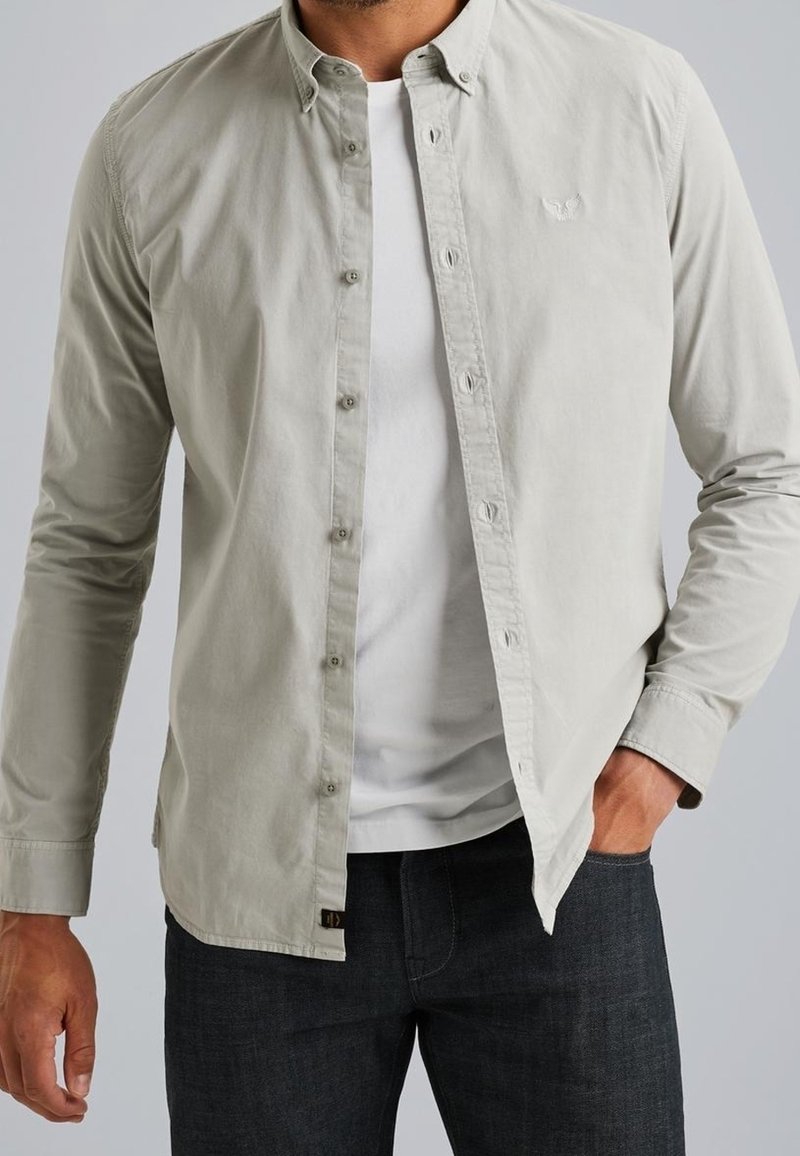 Homme portant une chemise gris clair à boutons ouverte sur un t-shirt blanc et un jean foncé, avec une main dans la poche, fond neutre.