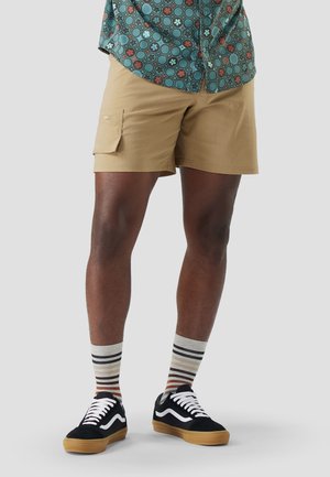 Person iført beige shorts med sidelomme, flerfarvede stribede sokker, sorte sneakers med hvide snørebånd og en grøn mønstret skjorte.