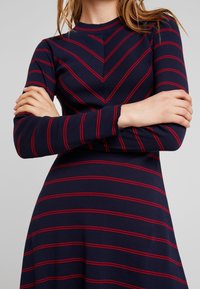 Abito blu navy a maniche lunghe con strisce orizzontali rosse, caratterizzato da un collo rotondo e un corpino aderente che si trasforma in una gonna a ruota.