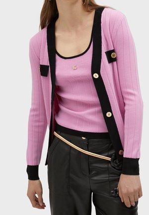 Žena na sobě má růžový žebrovaný cardigan a odpovídající tílko s černým lemováním, černé kalhoty a tenký zlatý řetízkový opasek.