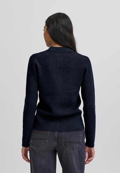 Jūras spēku džemperis ar pieguļošu dizainu un garām piedurknēm, izgatavots no mīksta materiāla. Ievērojama vienkārša apkakle un teksturēta apdare, saskaņots ar pelēkiem džinsiem.