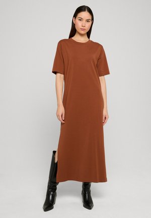 Femme debout, les mains détendues, portant une robe midi marron à manches courtes et des bottes noires mi-mollet, sur un fond clair uni.