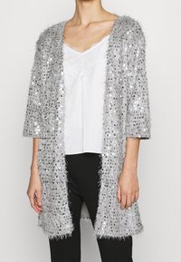 Sölsilver, lurvig cardigan med paljetter, öppen front och trekvartslånga ärmar, bärs över en vit tanktop och svarta byxor.