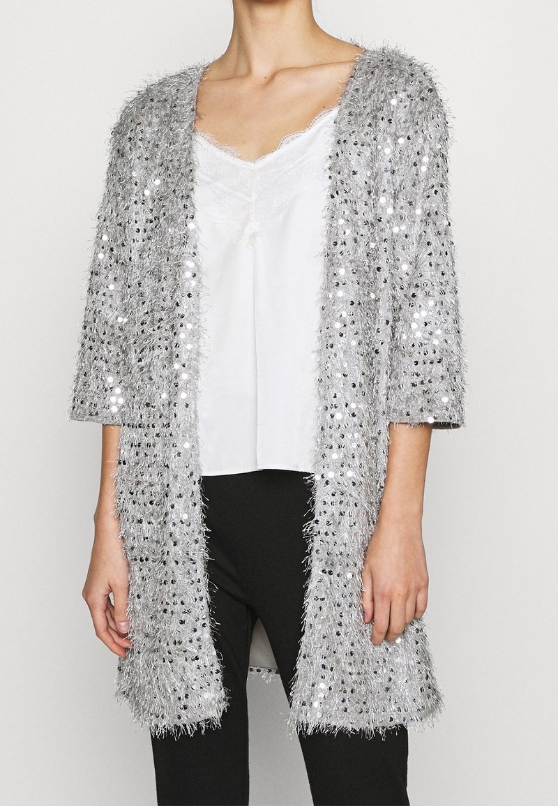 Sölsilver, lurvig cardigan med paljetter, öppen front och trekvartslånga ärmar, bärs över en vit tanktop och svarta byxor.