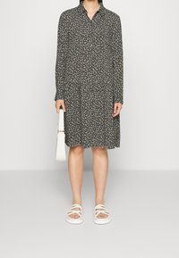 Robe noire avec motif floral blanc, manches longues, col avec boutons et jupe évasée. Associée à des sandales blanches et un sac à épaule blanc.
