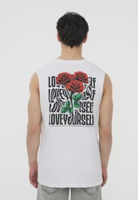 Maglietta bianca senza maniche con una grafica di rose rosse e testo nero che riporta frasi come "AMATI" sul retro.
