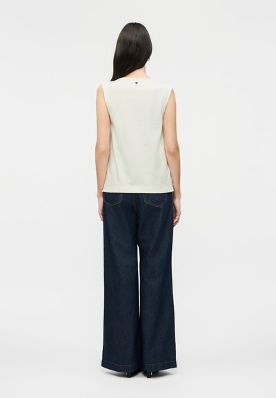 Γυναίκα που κοιτάζει προς τα πίσω, φοράει λευκό αμάνικο τοπ και σκούρα τζιν παντελόνια με φαρδιά μπατζάκια σε απλό φωτεινό φόντο.