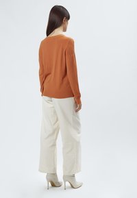 Orangenes Sweatshirt mit strukturierten Schultern, kombiniert mit weiten weißen Hosen und hellbeigen High-Heel-Stiefeln, vor einem schlichten Hintergrund.