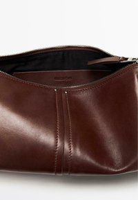 Pochette en cuir marron avec fermeture éclair argentée partiellement ouverte, révélant un intérieur en tissu noir et le nom de la marque embossé à l'intérieur.