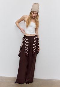 Hvid kort top, brede brune bukser og en beige strikket hue. Stribet sweater bundet om taljen. Glatte teksturer og neutrale farver.