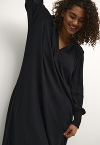 Black long-sleeve dress with a v-neckline, featuring a ribbed texture and flowing silhouette. Includes gathered accents at the sleeves.