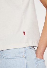 T-shirt en coton blanc avec une étiquette rouge Levi's sur la couture latérale, associé à un jean en denim bleu clair présentant un bouton argenté et une poche cousue.