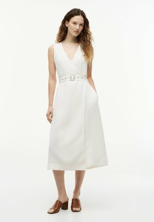 Femme aux longs cheveux bouclés portant une robe midi blanche sans manches avec ceinture et des sandales à talons carrés ouvertes marron, debout avec une main dans la poche.