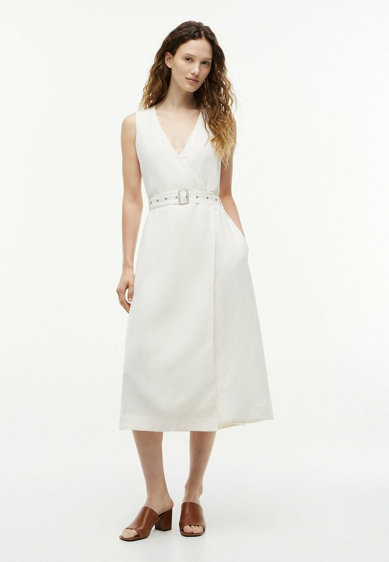 Femme aux longs cheveux bouclés portant une robe midi blanche sans manches avec ceinture et des sandales à talons carrés ouvertes marron, debout avec une main dans la poche.