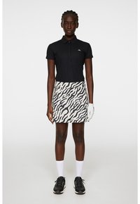 Svart pikétröja med textur och en liten logotyp, i kombination med en vit kjol med ett svart zebra-mönster. Modellen bär golfhandskar och skor.