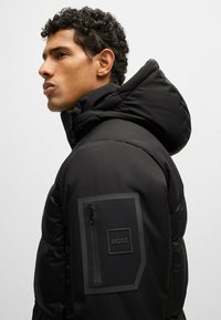 Black puffer jacket with a high collar and hood, featuring a zippered pocket on the left sleeve. Soft, smooth texture, and sleek design.