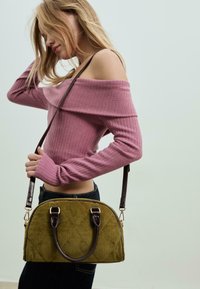 Olivově zelená kordurová taška s prošívaným vzorem, tmavohnědé kožené rukojeti a nastavitelný ramenní popruh. Modelka má na sobě růžový žebrovaný top.