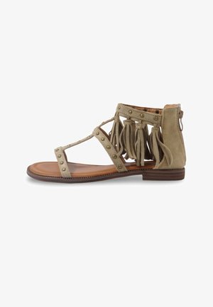 Sandale plate beige avec brides cloutées, détails en franges autour de la cheville, fermeture éclair à l'arrière et semelle marron.