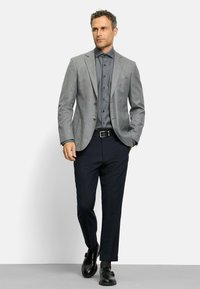 Grauer Wollblazer über einem dunklen Hemd mit Knöpfen, kombiniert mit dunklen Hosen und schwarzen Schuhen. Der Blazer hat zwei Knöpfe und einen Reverskragen.