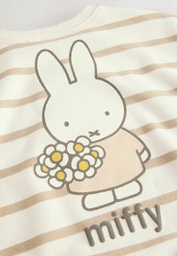 Bavlnený pulóver má béžové a krémové pruhy, s ilustráciou Miffy, ktorá drží kvety; text "miffy" je na spodnej strane s textúrou.