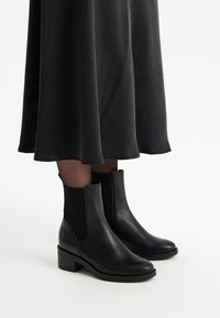 Czarne skórzane botki za kostkę z żebrowanymi panelami bocznymi, na słupkowym obcasie i gładkim wykończeniu. W połączeniu z czarną spódnicą, ukazującą fakturowany dół.