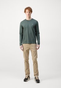 Langærmet grøn skjorte med rund hals, parret med beige bukser og sorte sneakers. Simpelt design uden synlige mønstre eller accenter.