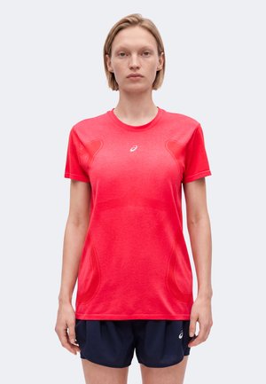 Mujer con el cabello rubio corto, que lleva una camiseta deportiva de manga corta de color rojo brillante y pantalones cortos de correr azul oscuro, de pie contra un fondo liso.